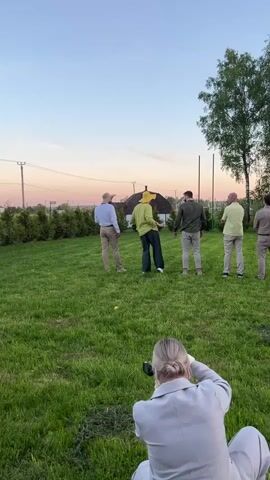 Here's a description of the video:

The video shows a group of men on a grassy lawn being photographed by a woman. The men are dressed in various shades of khaki, green, and white; one man wears a yellow hat and a matching overshirt. He appears to be dancing or performing for the photo. The other men are watching him. The woman is kneeling in the foreground, taking photos with her camera. In the background, there's a house, some bushes, and a tree. The sky is a light pink and blue, suggesting it might be sunset or sunrise. There are also power lines visible.