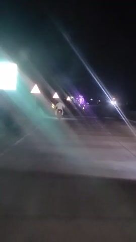 A person wearing a white garment is riding a motorcycle on a road at night. The motorcycle's taillight is illuminated, and dust or smoke is rising from the rear wheel. Several triangular warning signs with yellow borders are visible along the side of the road, illuminated by lights. In the distance, there are other lights, some appearing purple and white, suggesting other vehicles or structures. The sky is dark, indicating nighttime. The road appears to be paved.