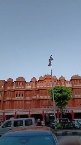 The video presents a ground-level view of a multi-story, red building with multiple domes on top, presumably a palace or historical building. The structure features intricate latticework and arched windows. On the ground floor, there are shops with visible signage in an Indian language. A streetlamp stands tall to the right of the building with two birds perched atop the lamp. In the foreground, portions of cars are visible parked along the street. The sky is clear and pale blue.