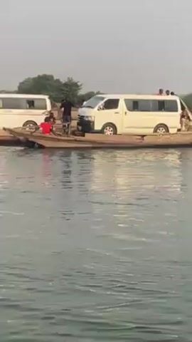 The video shows two white vans on a wooden raft or ferry in a body of water. Several people are on the raft, some standing near the vans. The ferry appears to be tilting. The surrounding environment consists of a sandy riverbank with some trees and buildings visible in the distance. The sky is overcast.