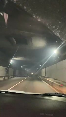The video captures a first-person perspective of driving through a tunnel. The interior of the car, specifically the dashboard and windshield wipers, are visible in the foreground. The tunnel itself is illuminated by artificial lights, casting a dim glow on the road and the tunnel walls. The road appears to be paved, with clear lane markings. There are other vehicles visible in the distance within the tunnel, indicated by their headlights. The tunnel walls are a light color, possibly concrete or painted. The overall atmosphere is somewhat dark and enclosed, typical of a tunnel.

The activity taking place is driving. The time of day is indeterminate due to the enclosed nature of the tunnel, but the artificial lighting suggests it could be nighttime or a particularly dark period. There are no obvious weather cues visible. Location cues are limited to what is typical of a tunnel on a road. No people are directly visible, but their presence is implied by the activity of driving. The mood could be described as functional or perhaps a bit somber due to the lack of natural light.

There is visible text on the dashboard, likely an airbag warning label, which reads "AIRBAG".