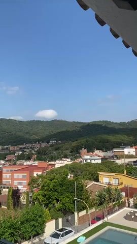 A wide shot pans from right to left across a sunlit residential area with a view of green, tree-covered hills under a bright blue sky with scattered white clouds. In the foreground, a swimming pool is visible on the right, with a white car parked on the left. A yellow house with arched windows and balconies is prominent in the middle ground. Behind it, several other houses with terracotta roofs and some apartment buildings are nestled among lush green trees. The camera movement is slow and smooth, offering a panoramic view of the urban landscape blending into the natural scenery. The lighting suggests it is daytime, likely late morning or early afternoon. No people are visible in the frame.
