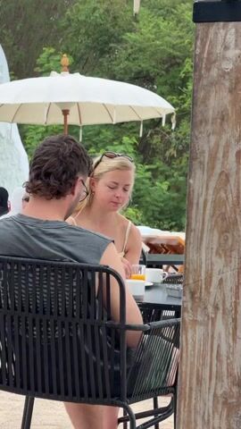 The video shows two separate scenes. The first scene features a couple sitting at a table outdoors, possibly at a restaurant or cafe. The man, seen from the back, has dark, curly hair and is wearing a grey sleeveless shirt. He is holding a phone to his ear. The woman, facing the camera, has blonde hair and is wearing a light-colored top. She is looking down, possibly at the table. On the table, there are drinks and possibly food. Behind them, a white umbrella is visible, and further back, greenery can be seen. The second scene shows a man in a red t-shirt and light-colored shorts taking a photo with his phone of a woman sitting at a table. The woman is wearing a red and green patterned dress and glasses, and she is smiling at the camera. She has a drink on the table in front of her. The background of this scene appears to be an outdoor setting with a railing and a cloudy sky.