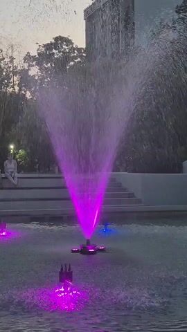 The video shows a fountain in a park at dusk. The fountain has colored lights that illuminate the water, creating a vibrant display. There is a woman sitting on a bench in the background, and trees and a building can be seen behind her. The water in the fountain is being sprayed in a fan-like pattern, and the lights are changing colors from purple to yellow and green.