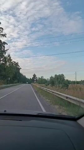 A forward-facing first-person perspective video shot from inside a car traveling on a paved road. The road curves to the left and is bordered by trees and vegetation. Overhead power lines are visible. The sky is blue with scattered clouds. A white van is seen driving ahead on the road. The dashboard of the car is visible in the foreground.