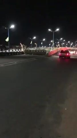 The video shows a street scene at night. The road is dark and paved. Streetlights line the road, illuminating the scene with a cool white light. There's a raised walkway or bridge with red accents, appearing to allow pedestrians to cross the road safely. A car with illuminated tail lights is seen driving on the road. On the left, there is a large truck or vehicle with a striped pattern along its side, possibly parked or stopped on the shoulder of the road. The sky is dark, indicating it's nighttime. There's minimal activity other than the car driving and the parked vehicle. The scene has a slightly grainy quality, common in nighttime video footage.