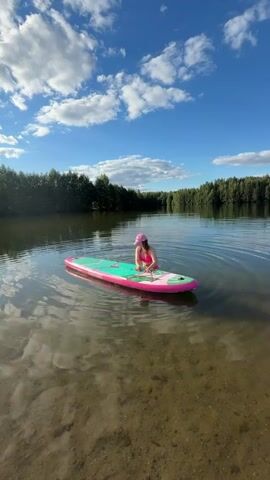 A person wearing a pink bikini and a pink baseball cap is sitting on a pink and green paddleboard in a lake. The person is reaching down and touching the paddleboard. The paddleboard is floating on the water. The background shows a forest and a blue sky with clouds. The water is calm and there are ripples on the surface. The sun is shining and casting reflections on the water.