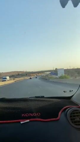 The video is filmed from the interior of a car driving on a highway. The dashboard mat, featuring the word "KONGO" in red stitching, is visible in the foreground. A blue sign with white Arabic script and the words "SUED TAFLOUT" is visible on the right side of the highway. Several cars and a truck are also seen on the road. A pedestrian bridge spans across the highway in the distance. The sky is clear and blue, suggesting it is daytime. The overall scene depicts a journey on a highway under a clear sky.