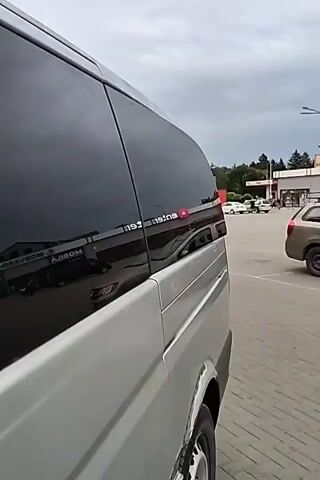The video shows the exterior of a silver van parked outdoors. The van has tinted windows and the reflection on the windows shows buildings and cars. The camera pans across the side of the van, showing the doors and windows. The environment appears to be a parking lot or a business area. There are no people visible in the video.