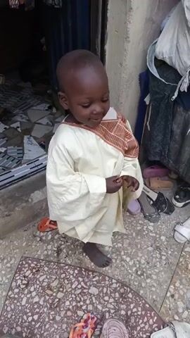 A young child dressed in a long-sleeved, off-white garment with brown embroidery on the shoulder area stands on a tiled surface. The child has dark skin, a bald head, and is barefoot. The child initially looks down, then turns towards the camera and smiles, before turning away again. Several pairs of shoes and sandals are scattered around the child on the tile floor. Behind the child is a closed blue metal door and a pile of fabric, possibly clothing. The tile surface is patterned with small, multi-colored fragments.