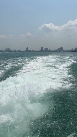 The video shows the wake of a boat moving through the water on a sunny day, with a distant city skyline visible on the horizon. The water is a dark turquoise color, churning into frothy white foam behind the boat. The sky is a hazy blue with scattered white clouds. The skyline consists of several buildings of varying heights, and what appears to be a construction crane is visible. The camera is positioned at the back of the boat.