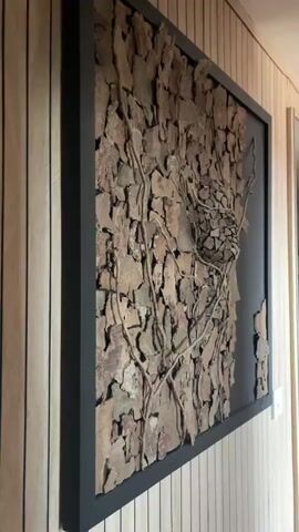 The video shows a close-up view of a framed piece of art made of natural materials. The art is mounted on a wall with vertical wooden planks. The artwork itself is a textured collage of irregular pieces of what appears to be bark or stone, arranged to form a rough, organic surface. Thin, twig-like branches are woven across the surface, creating a sense of natural growth. In the center of the artwork, there is a spherical or nest-like structure composed of smaller, fragmented pieces of bark or stone. The lighting casts shadows that emphasize the depth and texture of the artwork. The camera slowly pans across the artwork, providing different perspectives and highlighting its intricate details.