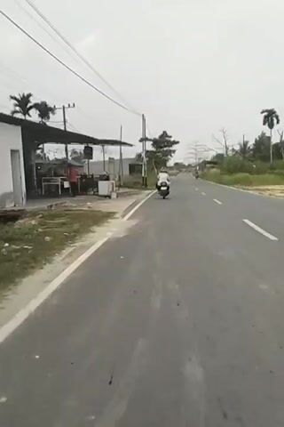 A motorcycle with a rider wearing a white top is traveling on a paved road. The rider is in the right lane of the road. The road is lined with grass and vegetation on the left side. There are some buildings visible on the left side of the road in the distance, including a structure with a covered area and some people standing near it. There are utility poles and wires overhead. The sky is overcast. The motorcycle is moving away from the camera. Another motorcycle is visible further down the road, also moving away from the camera.
