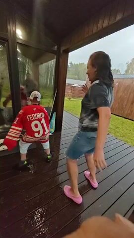 The video shows two people on a wet wooden deck. On the left, a child wearing a red hockey jersey with the name "FEDOROV" and the number "91" on the back is facing a glass door. The child is wearing a white baseball cap and dark shoes. On the right, an adult wearing a black and grey tie-dye t-shirt and denim shorts is facing away from the camera, towards the child. The adult is wearing pink slip-on shoes. The adult is holding a long, thin object, possibly a stick, and appears to be interacting with the child. The deck is wet, with water droplets visible. In the background, there is green grass, trees, and a wooden fence. The sky appears overcast, suggesting rain.