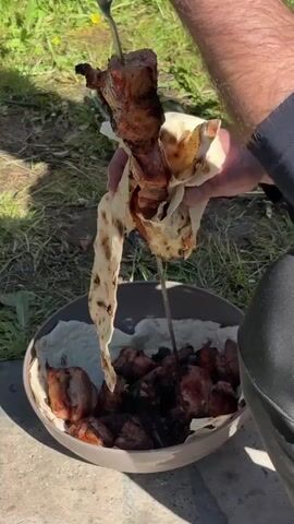 A person in dark pants and a black long sleeve shirt is holding a metal skewer of grilled meat, wrapped in flatbread, above a gray bowl filled with more grilled meat and flatbread. The bowl is sitting on the ground, among grass and dandelions. The person is pulling the meat off the skewer and into the bowl.