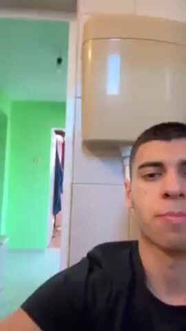 A young man is recording a video in what appears to be a bathroom. He is positioned in front of a beige toilet tank. The bathroom walls are tiled. Behind the man, partially visible through a doorway, is a room with a green wall. The man is wearing a black T-shirt. He gestures with his hand, licks his lips, and makes faces at the camera.
