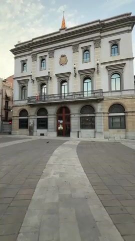 The video pans across a nearly empty town square with cobblestone paving. In the foreground, a white pathway curves through the square. Dominating the right side of the frame is a grand, ornate building with arched windows, a balcony, and a Spanish flag flying from the roof. The building appears to be a government or administrative building. To the left, several other multi-story buildings line the square, some with trees and awnings visible. The overall impression is one of quiet and stillness, possibly early morning or late evening.