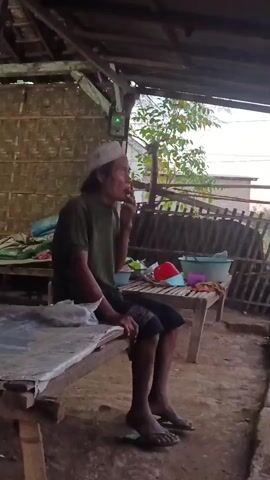 An elderly man wearing a white skullcap and a green t-shirt sits on a wooden bench. He has a cigarette in his mouth and is smoking. He is wearing dark shorts and flip-flops. The man is outdoors under a wooden shelter with a thatched roof. Behind him, there is a wall made of woven bamboo. To his right, on another bench, are several colorful plastic containers, including a red basin. The ground is dirt. In the background, there are trees and part of a building. The overall scene depicts a simple, rural setting.