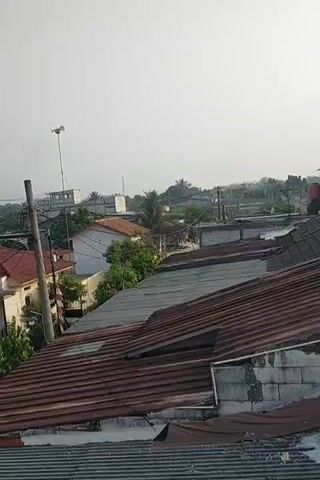 The video shows a neighborhood with several houses, mostly with corrugated metal roofs. The sky is overcast. There are trees and utility poles visible. In the foreground, there are roofs with some rust and damage. In the middle ground, there are houses with red tile roofs and some with white or light-colored walls. There are also some flags visible, including what appears to be an Indonesian flag. One house has a domed roof, possibly a mosque. There is a red object, possibly a car or boat, parked in front of one of the houses. A motorcycle is also parked nearby. In the background, there are more houses and trees. The overall impression is of a residential area.