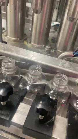 The video shows a close-up, eye-level view of an industrial liquid filling machine. Several clear plastic bottles are lined up and positioned under stainless steel filling nozzles. Black knob-like fixtures hold the bottles in place. As the video progresses, a yellow liquid begins to dispense from the nozzles into the bottles, filling them up. The background is blurred but appears to be part of the same industrial environment, with more stainless steel machinery. The lighting is bright and functional, typical of a manufacturing setting.