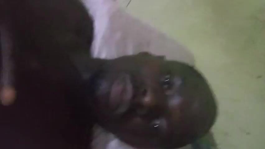 A dark-skinned man is lying down on a light-colored pillow. He is shirtless and his hand is resting on his chest. He is looking directly at the camera. The background is a light green wall.