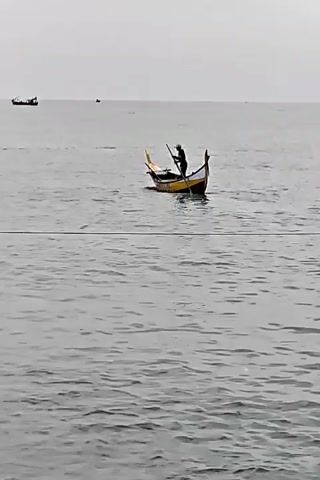 The video shows a person propelling a small, colorful boat through calm, gray water. The person stands in the center of the boat, using a long pole to push against the seabed, moving the boat forward. The boat has yellow and black sections, with decorative elements on the front. Other small boats are visible in the distance, and the sky is overcast, creating a flat, gray horizon.