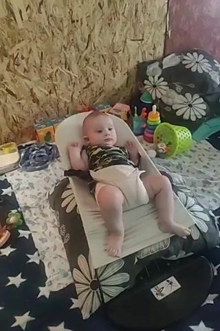 A baby is sitting in a white bouncer with a tan strap around its waist and legs. The baby is wearing a camouflage-print onesie and a diaper. The bouncer is on a bed with a blue and white star-patterned blanket. There are pillows with a black and white floral pattern on the bed. In the background, there is a wall made of wood panels. To the right of the baby, there is a stack of colorful plastic toys, including a ring stacker and a green basket. To the left of the baby, there are some small blocks. The lighting is dim.