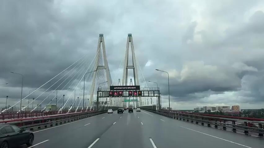 The video shows a view from a moving vehicle on a wet, multi-lane cable-stayed bridge. The sky is overcast and grey. The road has visible lane markings. Several cars are driving on the bridge. Ahead, there is a large overhead gantry with digital signs. The signs display traffic information and warnings, including speed limits of 90 and 70, road work warnings, and directional arrows. The bridge has two tall pylons with numerous cables extending from them. Streetlights are visible along the sides of the bridge. In the distance, buildings and more infrastructure can be seen. The overall impression is of a busy, urban bridge environment under cloudy weather conditions.