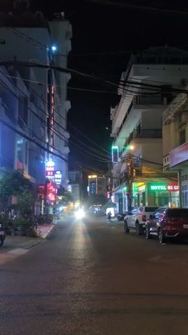 The video depicts a street scene at night. Buildings line both sides of the street, and numerous overhead wires are visible against the dark sky. The buildings are illuminated by a variety of neon signs in different colors, including green, blue, and red. Some signs advertise hotels and other establishments. Several parked cars and motorbikes are visible along the right side of the street. Car headlights shine brightly down the street, obscuring some details. A pedestrian is walking along the right side of the street, away from the camera. The street appears wet, reflecting the lights.