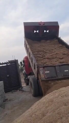 A red dump truck is backing up to a large pile of sand, and the truck bed is lifted up so the sand is pouring out of it onto the existing pile. The truck is parked on what appears to be a dirt lot near a brick wall and metal gate. The sky is overcast.