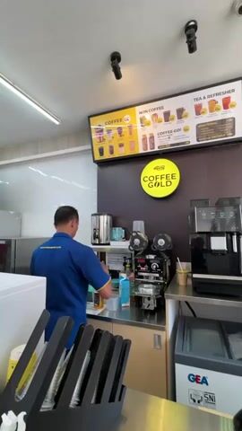 The video shows the interior of a coffee shop. A man in a blue shirt with a company logo on it is standing behind a counter, preparing a drink. He is holding a bottle of syrup and a dispenser. In front of him is a coffee machine and a cup sealing machine. To his left, there is a stainless steel counter and a refrigerator. Above the counter is a menu board displaying various coffee and tea options. The shop appears to be clean and well-lit.