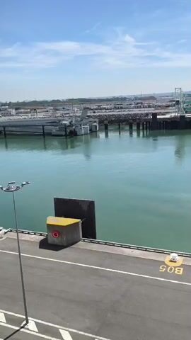 The video shows a view from a height overlooking a harbor on a sunny day. In the foreground, there is an asphalt area with a white arrow painted on it, and a lamppost on the left. Two concrete structures with yellow tops and a red ring are visible along the edge of the harbor. In the middle ground, there is a calm, turquoise body of water. Across the water, there is a pier with several pilings and a walkway. In the background, there is a cityscape with buildings and cranes, suggesting an industrial or port area. The sky is blue with some scattered white clouds. There are no people visible in the video.