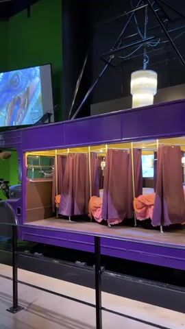 The video shows a purple tramcar with several booths, each with maroon curtains and some pink bedding. There are lights illuminating the interior of the tramcar, and a screen displaying an image of a dinosaur is visible in the background, to the left of the tramcar. A chandelier hangs from the ceiling above the tramcar. The tramcar is on a track, and the scene appears to be indoors, possibly in a museum or exhibition space. The video captures a voice counting down from three and then saying "yes."