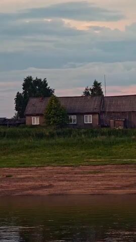 The video pans across a rural landscape by a body of water, likely a river, at dusk. The scene features a rustic village with several wooden houses and outbuildings situated on a grassy embankment overlooking a sandy shore. The houses appear aged, with weathered wood siding and dark-colored roofs. Some have visible windows, and one has a chimney. Trees and bushes are scattered around the buildings, adding to the natural setting.

In the foreground, the water surface is calm, reflecting the muted colors of the sky and the land. A sandy bank runs along the edge of the water, showing tire tracks, suggesting recent activity. A small, dark-colored motorboat is partially visible on the sandy shore, beached near the water's edge. Further along the bank, another, smaller boat or raft is also present.

The sky above is filled with clouds, painted with soft hues of pink, orange, and gray, indicative of either sunrise or sunset. The overall atmosphere is peaceful and serene, conveying a sense of quiet rural life. There is no visible text in the media file. The location is identified as Vorogovo, Russia.