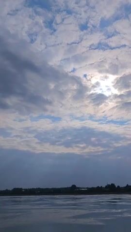 The video captures a wide shot of a body of water in the foreground, with a silhouetted treeline and buildings visible in the distance along the horizon. Above the treeline, the sky is filled with a mix of clouds. Some areas of the sky are clear blue, while others are covered by white and gray clouds. The sun is partially visible through a break in the clouds, creating a bright, hazy glow. The overall lighting suggests it is either sunrise or sunset. The water appears calm with subtle ripples reflecting the light from the sky. There are no people or discernible objects within the water itself, and no specific actions are taking place. The environment appears to be a natural landscape with human settlement in the background.