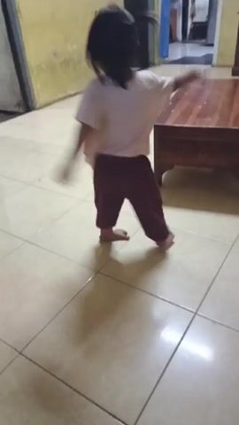 A young child with black hair in a ponytail and bangs is standing on a tiled floor. The child is wearing a white t-shirt with a Frozen print and maroon pants. In the background, there's a wooden coffee table and a doorway leading to another room. The walls are painted in a pale yellow color. The child is looking at the camera and appears to be posing or trying to imitate a photo model. Someone off-camera is encouraging the child to pose.