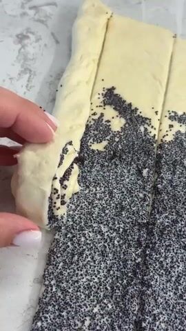 The video shows a close-up of a person's hands as they are preparing a pastry. The pastry dough is rolled out and covered with poppy seeds. The person is shown rolling up a section of the dough, revealing the poppy seed filling inside. The scene appears to be in a kitchen, with a marble-like countertop visible in the background. The lighting is bright and natural, suggesting daylight. The focus is on the detailed action of preparing the pastry.