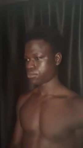 This is a close-up video of a dark-skinned male with a muscular build. He is shirtless and appears to be indoors, with a dark curtain in the background. The lighting is dim and highlights the contours of his chest and face. His head is turned to the left in the initial frames, and then he slowly turns to face the camera. His expression is neutral.