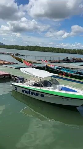 A white speed boat with green and blue accents is docked on a green floating platform. The boat has a white canopy and the word "muleara" is visible on its side in blue lettering. Several other longtail boats are docked behind it. The boats are in calm, green water. In the background, a line of trees forms the shoreline, and a few mountains are visible in the distance. The sky is blue with white, puffy clouds. The sun is shining.