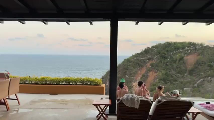 The camera shows a group of five people relaxing in a jacuzzi on a patio overlooking the ocean and a lush hillside. The patio has a covered area with a dining table and chairs. The jacuzzi is surrounded by lounge chairs. The ocean is visible in the distance under a sky with pastel colors suggesting dusk. The group appears to be talking and enjoying their time together.
