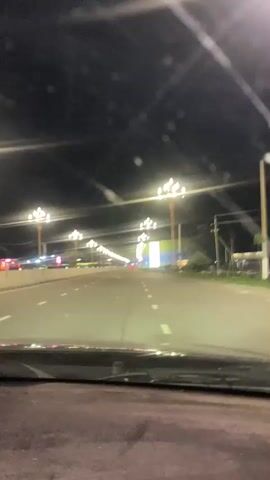 The video shows a first-person perspective from inside a vehicle driving on a road at night. The road is lit by streetlights on tall poles. There are other vehicles visible on the road, and some structures and billboards along the side. The scene suggests a suburban or urban environment.