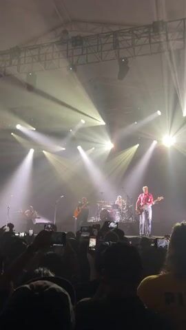 A band performs on a stage in front of a crowd. The band consists of at least five members: a keyboard player on the left, two guitarists in the middle, a drummer in the back, and a bassist on the right.  The bassist is wearing a red shirt and jeans. The stage is lit by many white spotlights and filled with smoke. The audience is holding up their phones to record the performance. There is a large metal lighting rig above the stage. The event appears to be held inside a large tent or building.