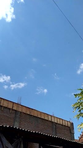A video clip looking up at the sky. A clear blue sky with some white clouds is visible. In the foreground, there is the edge of a building under construction. The building has brick walls and wooden framing. Some leaves from a tree are visible at the top edge of the frame.