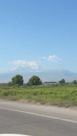 A video recorded from a moving vehicle shows a view of Mount Ararat, a snow-capped mountain range, on a clear day. The foreground of the video shows a road, a patch of green grass, and some trees. The background features the mountain range, with clouds partially obscuring its peak. The sky is a clear blue. There are no people or other significant objects visible in the video. The video appears to be a travelogue or scenic footage.