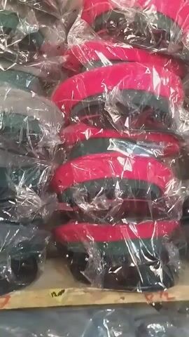 The video shows stacks of berets wrapped in clear plastic. Some berets are red and green, while others are dark green. The berets are piled on wooden pallets. The lighting is dim, and the background is blurry.
