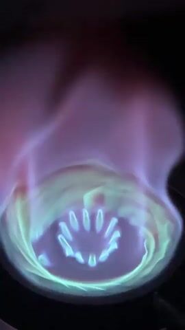 The video captures a close-up of a gas stove burner with active flames in a dark setting, likely at nighttime or in a dimly lit room. The flames display a vibrant mix of colors: intense blue at the base, transitioning into a prominent, swirling green in the middle section, and fading into a pinkish-purple hue at the top. The individual gas outlets of the burner are visible, glowing blue within the flame's core. No other objects or people are visible in the frame, and there is no visible text. The location context provided is Angwan Sayawa, Nigeria.