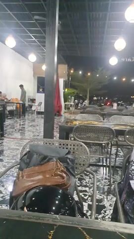 The image depicts a nighttime scene at a casual outdoor or semi-outdoor restaurant or cafe in Glameunasah Baro, Indonesia.

In the immediate foreground, a light brown leather handbag, a black backpack, and a glossy black motorcycle helmet are placed on a woven metal chair. This chair is pulled up to a dark-surfaced table, possibly marble. To the right, part of another chair with clothing draped over it is visible.

The midground features several empty tables with dark surfaces and matching woven metal chairs, arranged in rows. Black structural pillars support the roof, from which multiple spherical white light fixtures hang, illuminating the space. The floor appears to have a dark, possibly marbled or patterned, finish.

In the background, to the left, a man in a light green shirt stands near a counter or service area, possibly waiting to order or for food. Further back and outside the main covered area, more artificial lights are visible, along with glimpses of trees and what appear to be parked motorcycles or scooters, indicating customers' transportation. On the far right wall, mirrored text is visible, appearing to read "916 KAFE" or a similar establishment name.

The overall atmosphere is calm and relaxed, suggesting a casual dining or social gathering spot during the evening. No specific activity beyond waiting or preparing to dine is clearly depicted, but the setup implies a typical restaurant environment.
