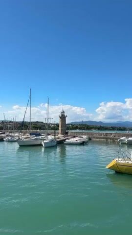 The video shows a harbor on a clear day. The water is a vibrant turquoise color, and the sky is a brilliant blue with scattered white clouds. Several sailboats are docked in the harbor, their masts reaching towards the sky. In the background, there is a lighthouse, and beyond that, a line of hills or mountains. The overall impression is one of a peaceful and scenic maritime setting.