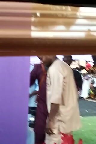 The video shows two people standing next to each other. The person on the right is wearing a white outfit, while the person on the left is wearing a tan outfit. The video is taken from a low angle, and there are flowers in the foreground. The background appears to be a gathering of people. The video is shaky and the quality is not great.