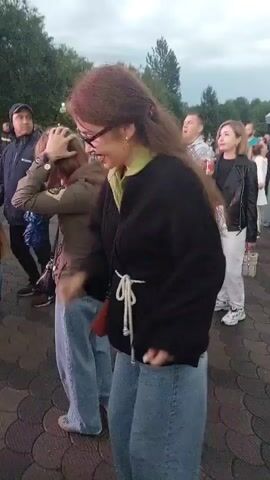 The video shows an outdoor gathering of people. In the foreground, a young woman with long brown hair and glasses is dancing. She is wearing a black jacket over a green shirt and blue jeans. To her left, another woman with short reddish-brown hair is also dancing and holding a child. Behind them, several other people are visible, some looking towards the front, others interacting with each other. The ground appears to be paved with cobblestones, and there are trees and foliage in the background. The sky is overcast, suggesting it might be late in the day or cloudy weather. The overall atmosphere appears to be casual and festive.